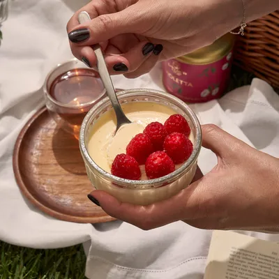 Dettaglio di una mano che aggiunge lamponi freschi sopra un dessert cremoso servito in una ciotola su vassoio.