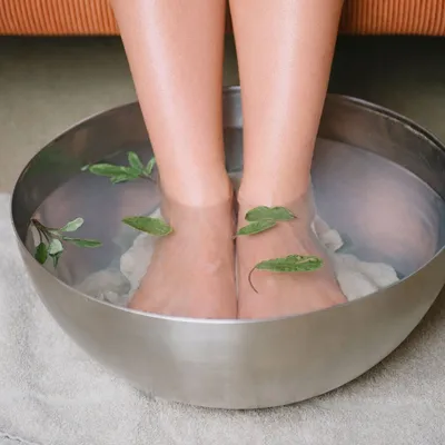 Gambe immerse fino alle caviglie in una bacinella metallica per pediluvio, con foglie o erbe aromatiche che galleggiano nell’acqua.