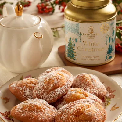 Piatto di dolci rotondi spolverati di zucchero a velo, con barattolo di tè e teiera sfocati sullo sfondo.