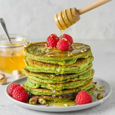 Torre di pancake verdi, probabilmente al matcha, con miele che cola dall’alto e lamponi freschi come decorazione.