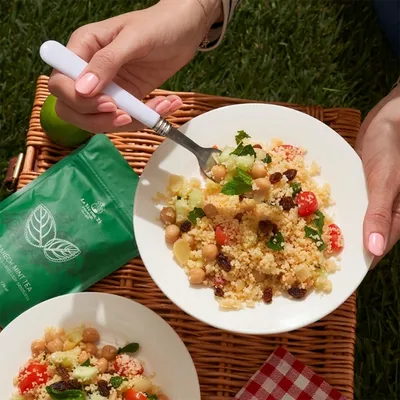 Scena di picnic all’aperto con picnic con piatto di pasta e borsa verde, piatti e accessori disposti su una tovaglia o sul prato.
