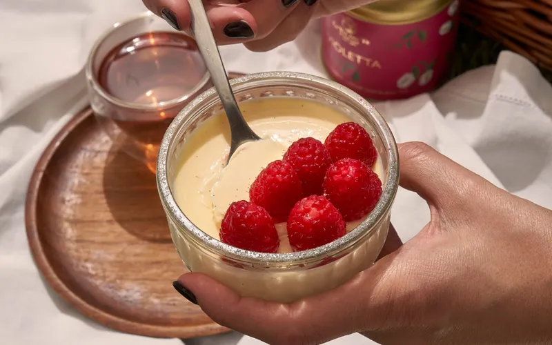 Dettaglio di una mano che aggiunge lamponi freschi sopra un dessert cremoso servito in una ciotola su vassoio.