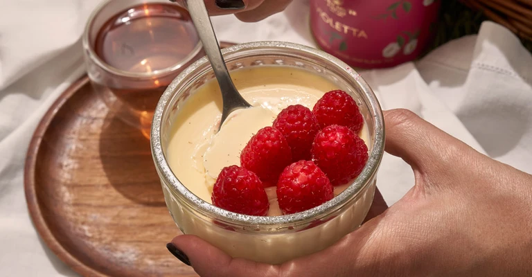 Détail d'une main qui aggiunge framboises frais sopra un dessert crémeux servito dans une bol sur plateau.
