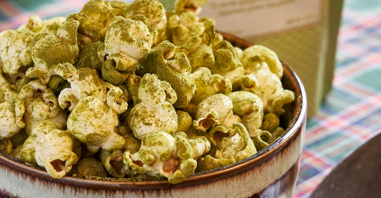 Primo piano di ciotola di popcorn verdi al matcha, impiattato con cura su un tavolo o sfondo neutro, con ingredienti e decorazioni visibili attorno.