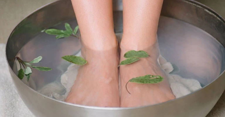 Gambe immerse fino alle caviglie in una bacinella metallica per pediluvio, con foglie o erbe aromatiche che galleggiano nell’acqua.