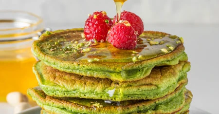 Torre di pancake verdi, probabilmente al matcha, con miele che cola dall’alto e lamponi freschi come decorazione.